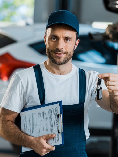 happy car mechanic looking at camera and holding clipboard and key near car